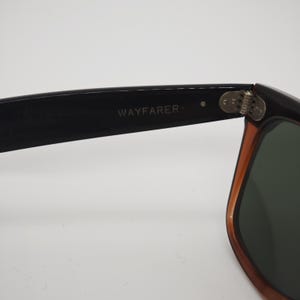 May include: Close-up of Wayfarer sunglasses. The black frame transitions to a brown hue near the lenses. The text "WAYFARER" is printed on the black arm of the sunglasses. The lenses are a dark green color.