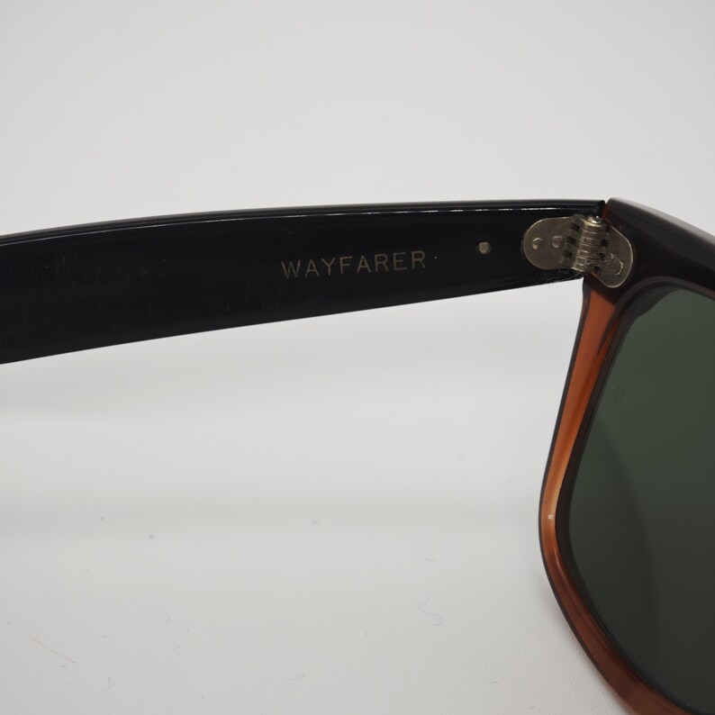 May include: Close-up of Wayfarer sunglasses. The black frame transitions to a brown hue near the lenses. The text "WAYFARER" is printed on the black arm of the sunglasses. The lenses are a dark green color.