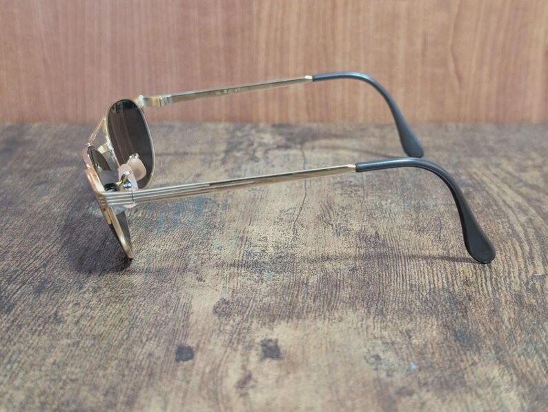 May include: Gold-tone aviator sunglasses with dark lenses. The sunglasses have a classic teardrop shape, a thin gold metal frame, and black temple tips. The lenses appear to be tinted, offering a sleek, vintage aesthetic.
