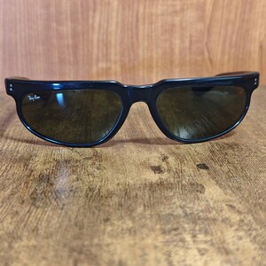 May include: Black sunglasses with dark lenses. The sunglasses have a thick, curved frame with a subtle design on the temples. The Ray-Ban logo is visible on the left lens. The sunglasses are resting on a wooden surface.