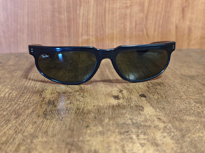 May include: Black sunglasses with dark lenses. The sunglasses have a thick, curved frame with a subtle design on the temples. The Ray-Ban logo is visible on the left lens. The sunglasses are resting on a wooden surface.
