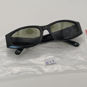 Może przedstawiać: Czarne okulary przeciwsłoneczne z prostokątną oprawką. Logo Ray-Ban jest widoczne na lewej soczewce. Soczewki są w ciemnozielonym kolorze. Okulary przeciwsłoneczne leżą na białej powierzchni.