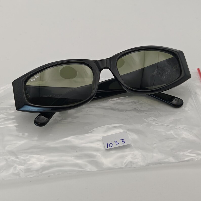 Może przedstawiać: Czarne okulary przeciwsłoneczne z prostokątną oprawką. Logo Ray-Ban jest widoczne na lewej soczewce. Soczewki są w ciemnozielonym kolorze. Okulary przeciwsłoneczne leżą na białej powierzchni.