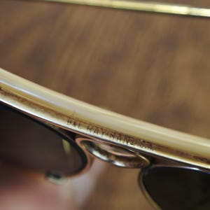 May include: Close-up of vintage B&L Ray-Ban sunglasses. The frame is a light cream color with gold accents. The lenses are dark, and the text "B&L RAY-BAN U.S.A." is visible on the frame.