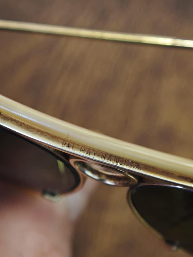 May include: Close-up of vintage B&L Ray-Ban sunglasses. The frame is a light cream color with gold accents. The lenses are dark, and the text "B&L RAY-BAN U.S.A." is visible on the frame.