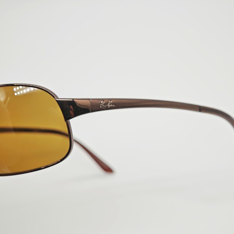 May include: Brown sunglasses with amber-colored lenses. The sunglasses have a sleek, modern design with a thin frame and curved temples. The Ray-Ban logo is visible on the temple.