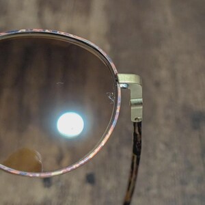 Peut inclure: Gros plan de lunettes de soleil rondes avec une monture &agrave; motifs dans les tons rose, orange et bleu. Les verres sont bruns et les branches ont un imprim&eacute; animalier marron et noir. La monture est dor&eacute;e.