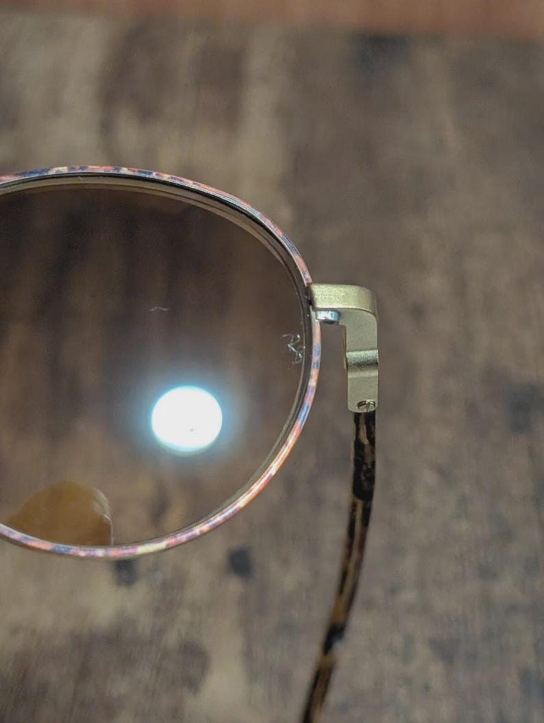 Peut inclure: Gros plan de lunettes de soleil rondes avec une monture &agrave; motifs dans les tons rose, orange et bleu. Les verres sont bruns et les branches ont un imprim&eacute; animalier marron et noir. La monture est dor&eacute;e.