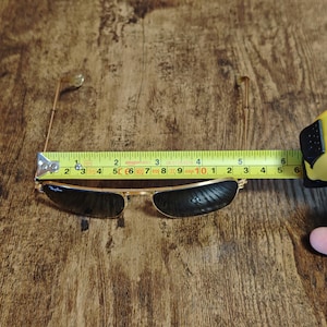 Puede incluir: Gafas de sol con montura dorada y lentes oscuras, de aproximadamente 12,7 cm de ancho. Las gafas est&aacute;n sobre una superficie de madera, con una cinta m&eacute;trica. El logotipo de Ray-Ban es visible en la lente.