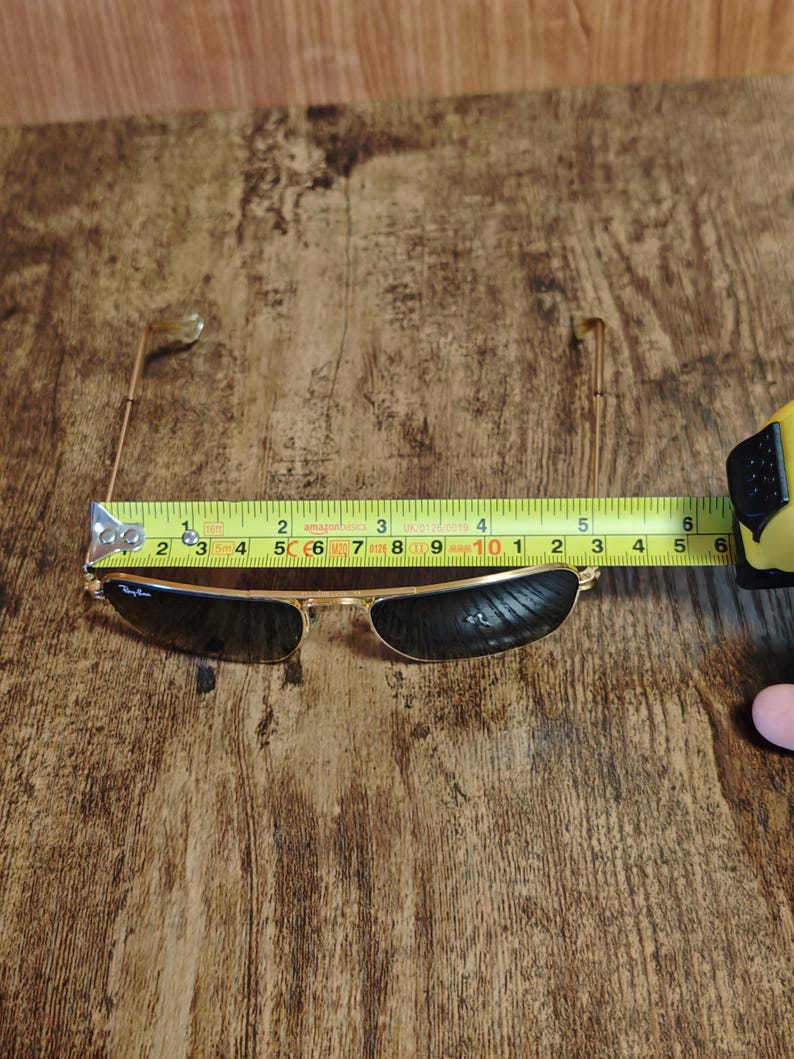 Puede incluir: Gafas de sol con montura dorada y lentes oscuras, de aproximadamente 12,7 cm de ancho. Las gafas est&aacute;n sobre una superficie de madera, con una cinta m&eacute;trica. El logotipo de Ray-Ban es visible en la lente.