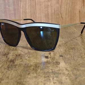 Peut inclure: Lunettes de soleil noires avec une monture en m&eacute;tal dor&eacute; et des verres fonc&eacute;s. Les lunettes ont une forme &eacute;paisse et rectangulaire avec un accent dor&eacute; sur le dessus de la monture. Le logo Ray-Ban est visible.