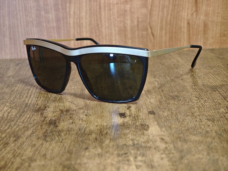 Peut inclure: Lunettes de soleil noires avec une monture en m&eacute;tal dor&eacute; et des verres fonc&eacute;s. Les lunettes ont une forme &eacute;paisse et rectangulaire avec un accent dor&eacute; sur le dessus de la monture. Le logo Ray-Ban est visible.