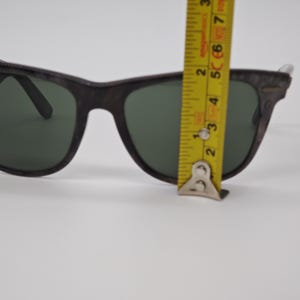 Puede incluir: Gafas de sol marrones oscuras con lentes verdes. Se miden con una cinta m&eacute;trica amarilla. La montura tiene un aspecto texturizado. Las lentes son de color verde oscuro.