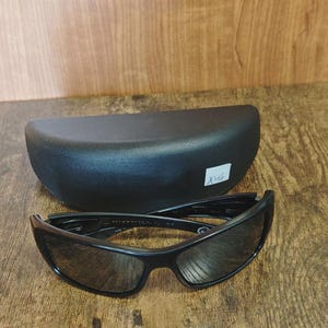 K&ouml;nnte beinhalten: Schwarze Sonnenbrille mit passendem schwarzem Etui. Die Sonnenbrille hat ein schlankes, modernes Design mit dunklen Gl&auml;sern und einem stabilen Rahmen. Das ovale Etui bietet Schutz f&uuml;r die Brille.