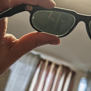 May include: Black sunglasses with dark green lenses are held in a hand. The sunglasses have a thick, rectangular frame. The lenses reflect the surrounding environment. The arms of the sunglasses are black and have a silver emblem.