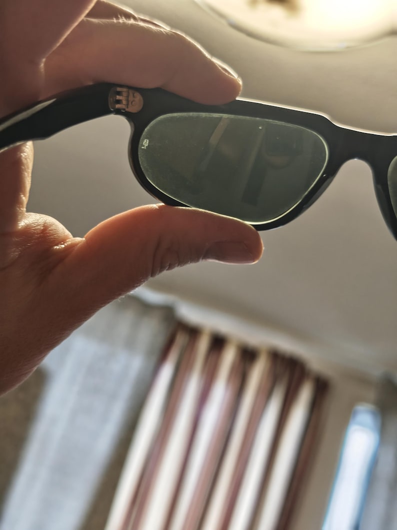 May include: Black sunglasses with dark green lenses are held in a hand. The sunglasses have a thick, rectangular frame. The lenses reflect the surrounding environment. The arms of the sunglasses are black and have a silver emblem.