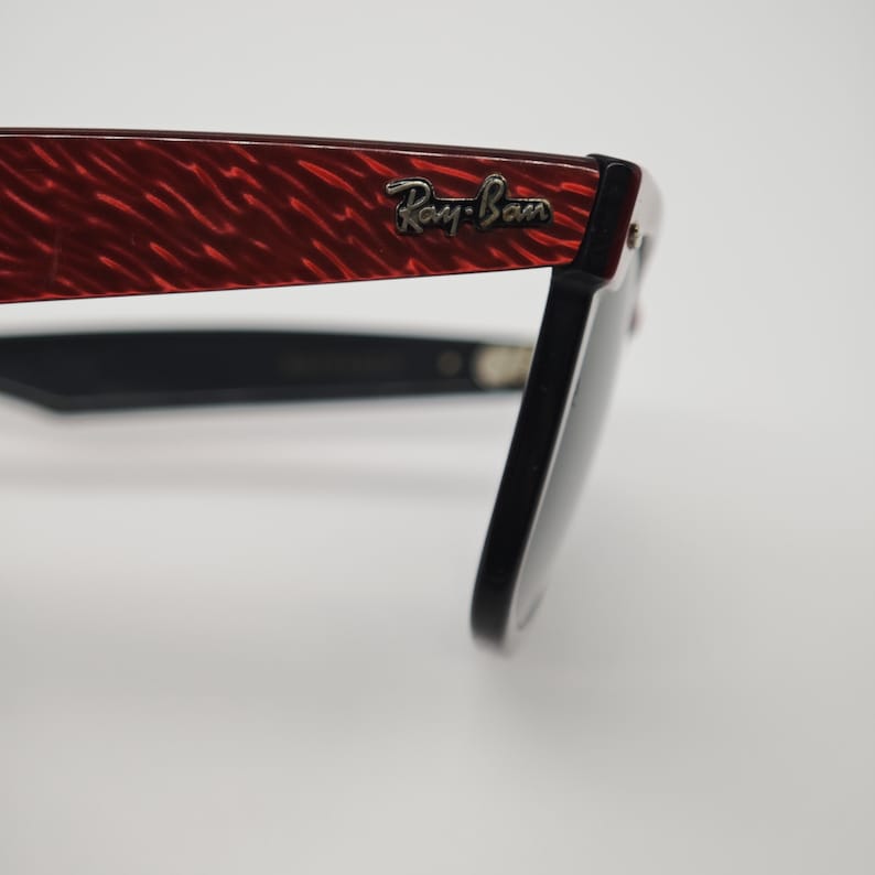 May include: Close-up of Ray-Ban sunglasses. The frame has a red, textured pattern on the temples, with the Ray-Ban logo in silver. The lenses are black, and the arms are black.