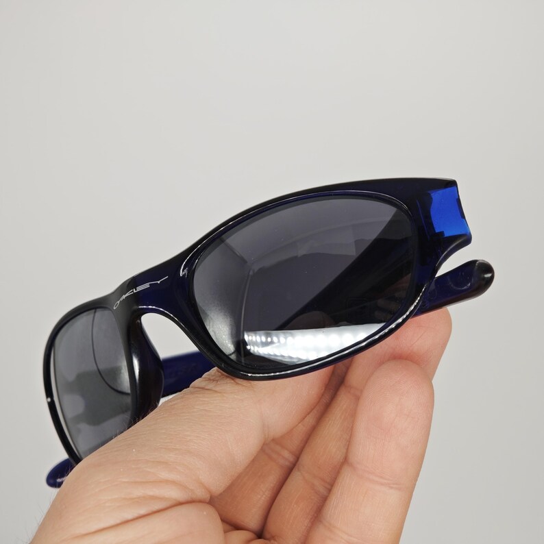 May include: Dark blue sunglasses with a curved design. The lenses are dark, and the frame has a glossy finish with blue accents. The brand name is visible on the frame.