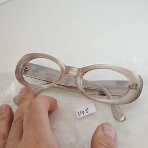 May include: Gianni Versace eyeglasses with oval-shaped, translucent beige frames. The inner temple arm features the text GIANNI VERSACE MADE IN ITALY. The glasses are positioned on a clear plastic surface with a small white tag labeled 198.