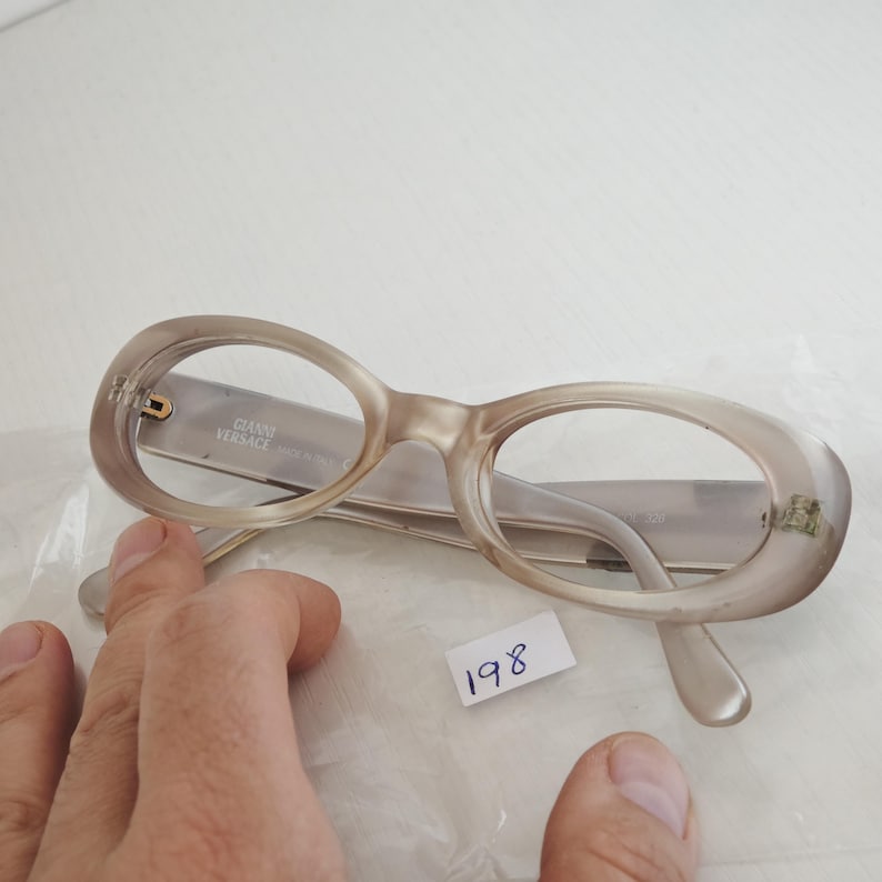 May include: Gianni Versace eyeglasses with oval-shaped, translucent beige frames. The inner temple arm features the text GIANNI VERSACE MADE IN ITALY. The glasses are positioned on a clear plastic surface with a small white tag labeled 198.