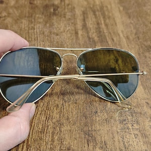 May include: Aviator-style sunglasses with a gold-colored metal frame and dark lenses. The sunglasses are held by a hand, showcasing the classic design and shape. The lenses have a slight green tint.
