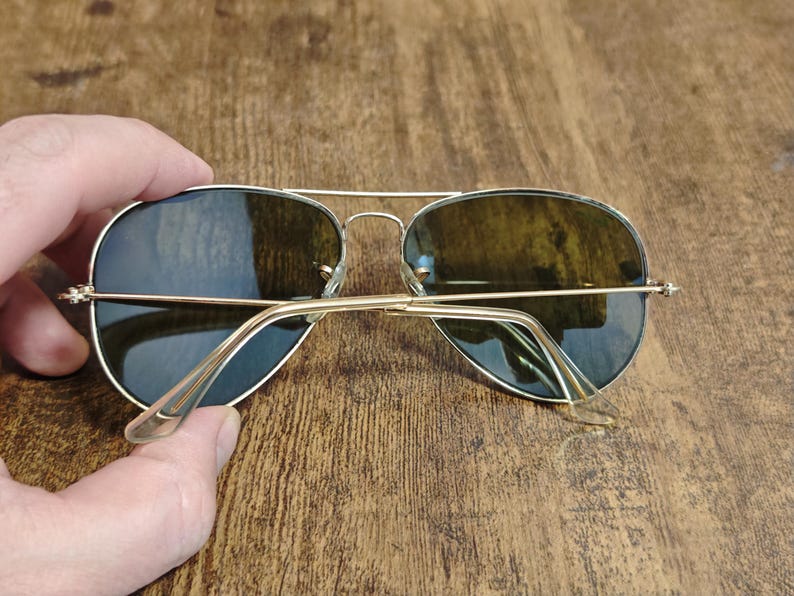 May include: Aviator-style sunglasses with a gold-colored metal frame and dark lenses. The sunglasses are held by a hand, showcasing the classic design and shape. The lenses have a slight green tint.