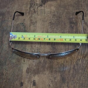 May include: Sunglasses with a silver frame and brown lenses are measured with a tape measure. The sunglasses measure approximately 5 inches in width. The arms of the sunglasses are black.