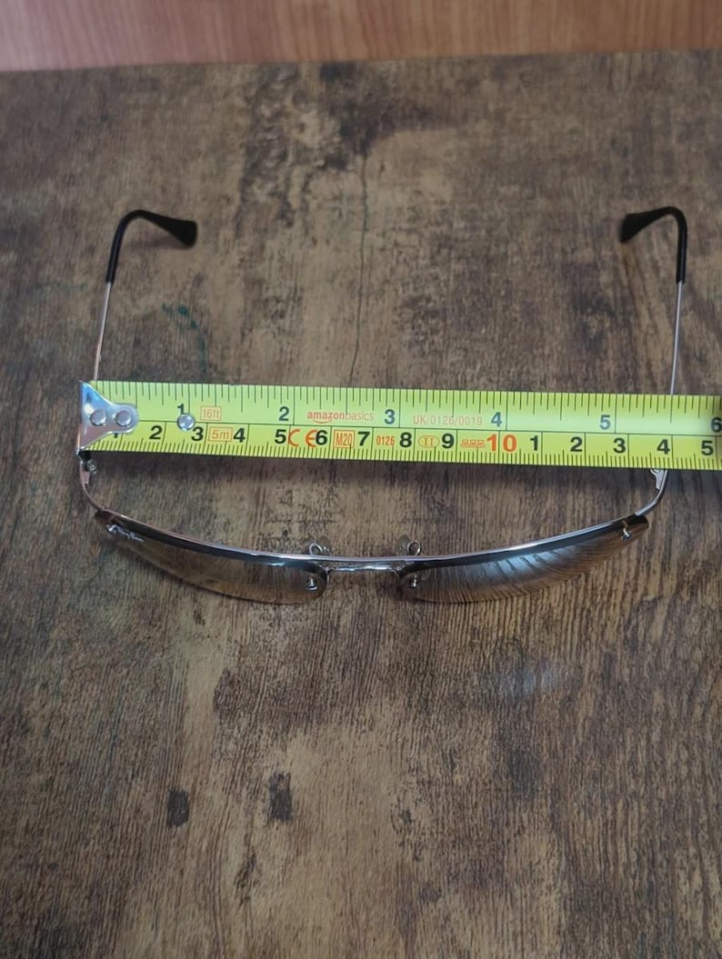 May include: Sunglasses with a silver frame and brown lenses are measured with a tape measure. The sunglasses measure approximately 5 inches in width. The arms of the sunglasses are black.