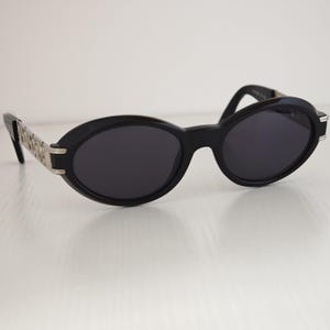 Puede incluir: Gafas de sol de forma ovalada con montura negra y lentes oscuras. Las patillas presentan un dise&ntilde;o de eslabones met&aacute;licos en tono plateado que se conecta con la montura frontal. El puente est&aacute; integrado en la estructura de pl&aacute;stico negro.