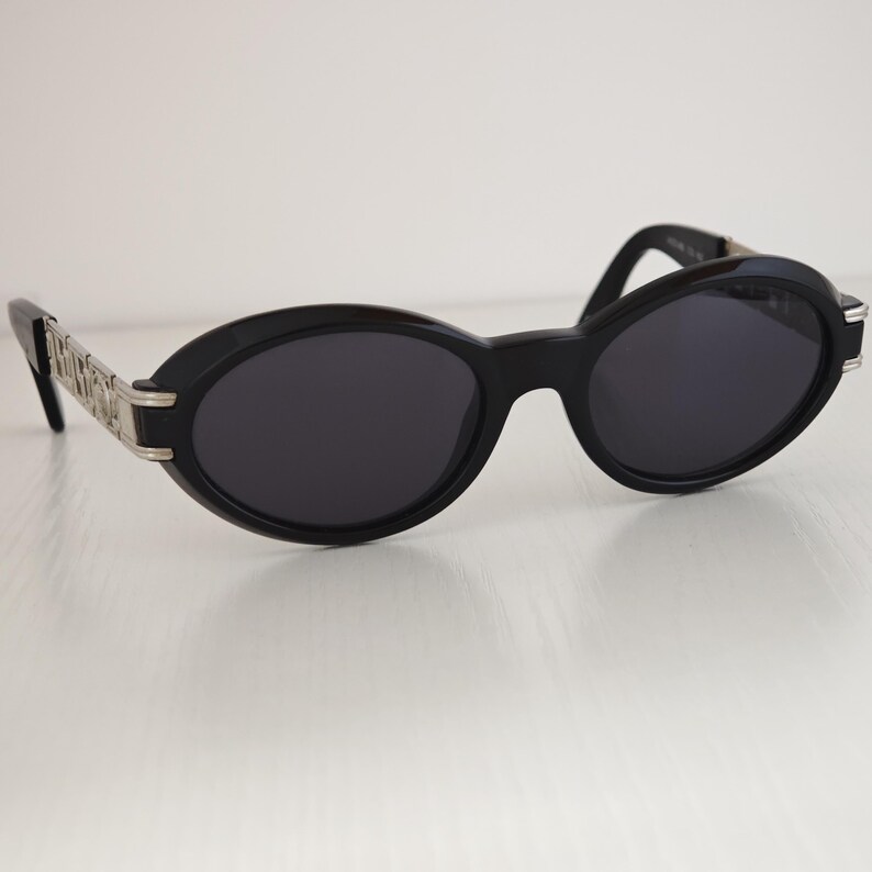 Puede incluir: Gafas de sol de forma ovalada con montura negra y lentes oscuras. Las patillas presentan un dise&ntilde;o de eslabones met&aacute;licos en tono plateado que se conecta con la montura frontal. El puente est&aacute; integrado en la estructura de pl&aacute;stico negro.