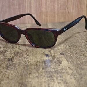 May include: Sunglasses with a tortoiseshell pattern frame and dark lenses. The arms are black with a logo on the side. The sunglasses are resting on a wooden surface.