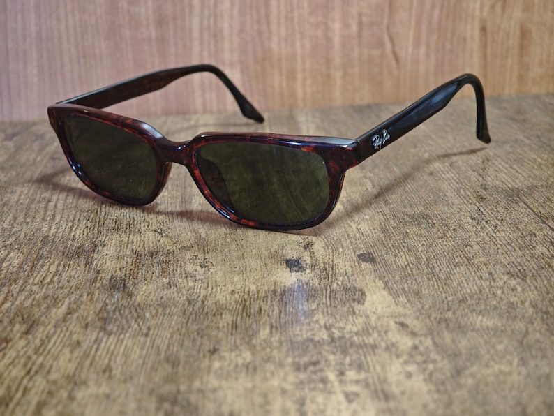 May include: Sunglasses with a tortoiseshell pattern frame and dark lenses. The arms are black with a logo on the side. The sunglasses are resting on a wooden surface.