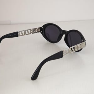 Puede incluir: Gafas de sol ovaladas negras con lentes oscuras y patillas de metal plateado con un patr&oacute;n geom&eacute;trico recortado. El brazo interior lleva el texto MOD 486 COL 462. El dise&ntilde;o incluye un emblema circular plateado en la bisagra de la patilla.