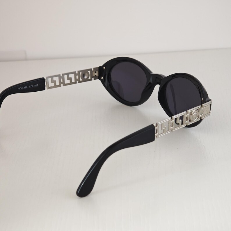 Puede incluir: Gafas de sol ovaladas negras con lentes oscuras y patillas de metal plateado con un patr&oacute;n geom&eacute;trico recortado. El brazo interior lleva el texto MOD 486 COL 462. El dise&ntilde;o incluye un emblema circular plateado en la bisagra de la patilla.