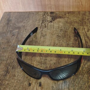 K&ouml;nnte beinhalten: Schwarze Sonnenbrille mit geschwungenem Design, dunklen Gl&auml;sern und schwarzem Rahmen. Ein Ma&szlig;band liegt &uuml;ber der Sonnenbrille und zeigt eine L&auml;nge von etwa 15 cm.