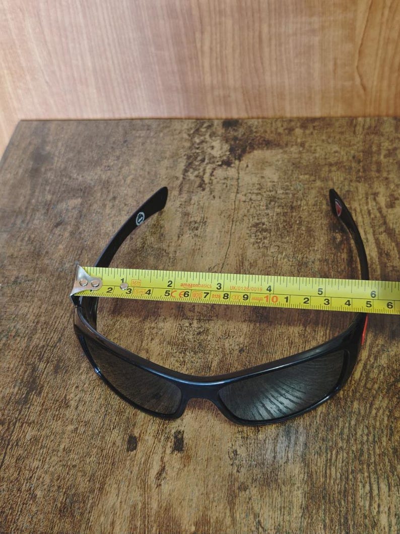 K&ouml;nnte beinhalten: Schwarze Sonnenbrille mit geschwungenem Design, dunklen Gl&auml;sern und schwarzem Rahmen. Ein Ma&szlig;band liegt &uuml;ber der Sonnenbrille und zeigt eine L&auml;nge von etwa 15 cm.
