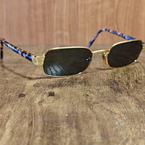 May include: Gold-framed sunglasses with dark lenses. The temples feature a blue and brown tortoiseshell pattern. The sunglasses are rectangular and rest on a wooden surface.