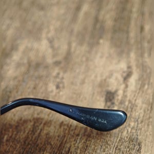 Peut inclure: Gros plan d'une branche de lunettes noire avec le texte "Ray-Ban USA" imprim&eacute; en blanc. La branche est incurv&eacute;e et a une finition brillante, sur un fond en bois flou.