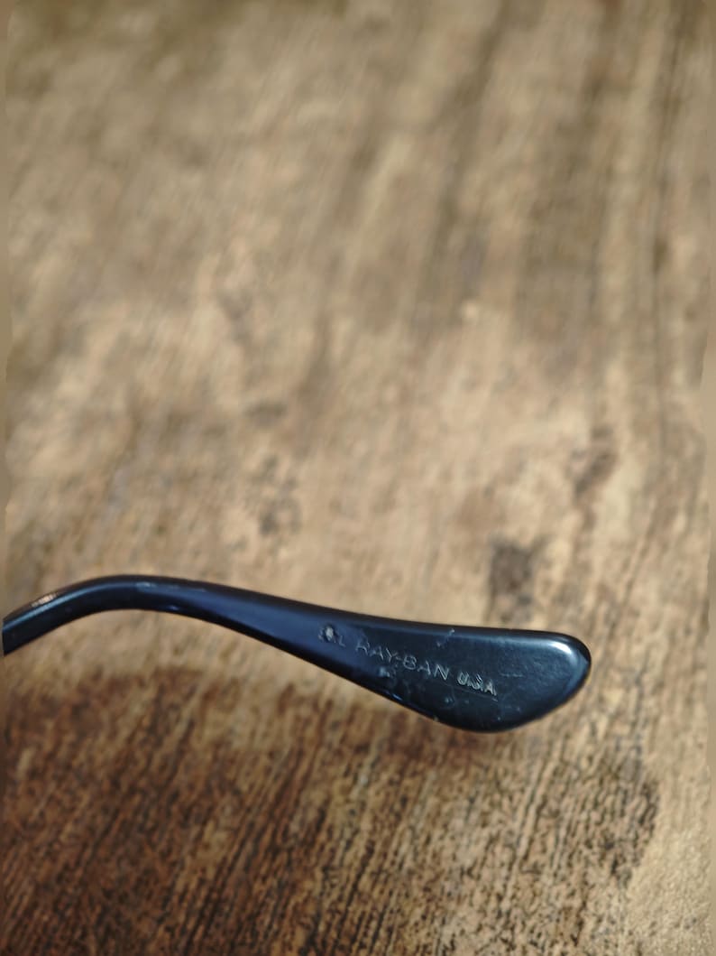 Peut inclure: Gros plan d'une branche de lunettes noire avec le texte "Ray-Ban USA" imprim&eacute; en blanc. La branche est incurv&eacute;e et a une finition brillante, sur un fond en bois flou.