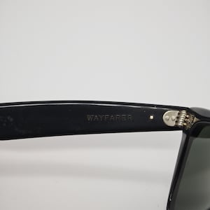 May include: Close-up of black Wayfarer sunglasses. The text "WAYFARER" is visible on the temple arm. The sunglasses have a classic design with a dark frame and tinted lenses. The hinge is silver.