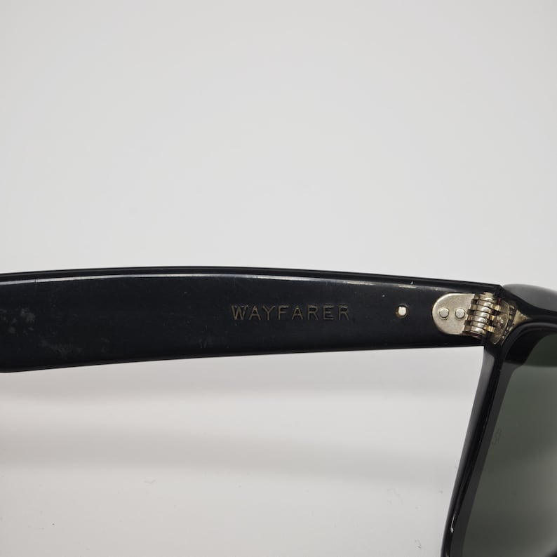 May include: Close-up of black Wayfarer sunglasses. The text "WAYFARER" is visible on the temple arm. The sunglasses have a classic design with a dark frame and tinted lenses. The hinge is silver.