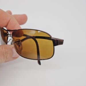 May include: Brown-framed sunglasses with amber-tinted lenses. The rectangular lenses are set in a thin, dark brown metal frame. The temples are also dark brown. The sunglasses are held against a white background.