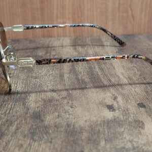 Peut inclure: Lunettes de soleil &agrave; monture dor&eacute;e avec verres bruns. Les branches pr&eacute;sentent un motif &agrave; motifs marron, beige et noir. Les lunettes sont pos&eacute;es sur une surface en bois, montrant le profil lat&eacute;ral.