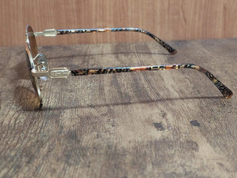 Peut inclure: Lunettes de soleil &agrave; monture dor&eacute;e avec verres bruns. Les branches pr&eacute;sentent un motif &agrave; motifs marron, beige et noir. Les lunettes sont pos&eacute;es sur une surface en bois, montrant le profil lat&eacute;ral.