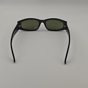 Może przedstawiać: Czarne okulary przeciwsłoneczne z prostokątnymi oprawkami i zielonymi szkłami. Okulary mają elegancki, nowoczesny design z lekko zakrzywioną oprawką i zausznikami. Szkła mają stonowany zielony kolor.