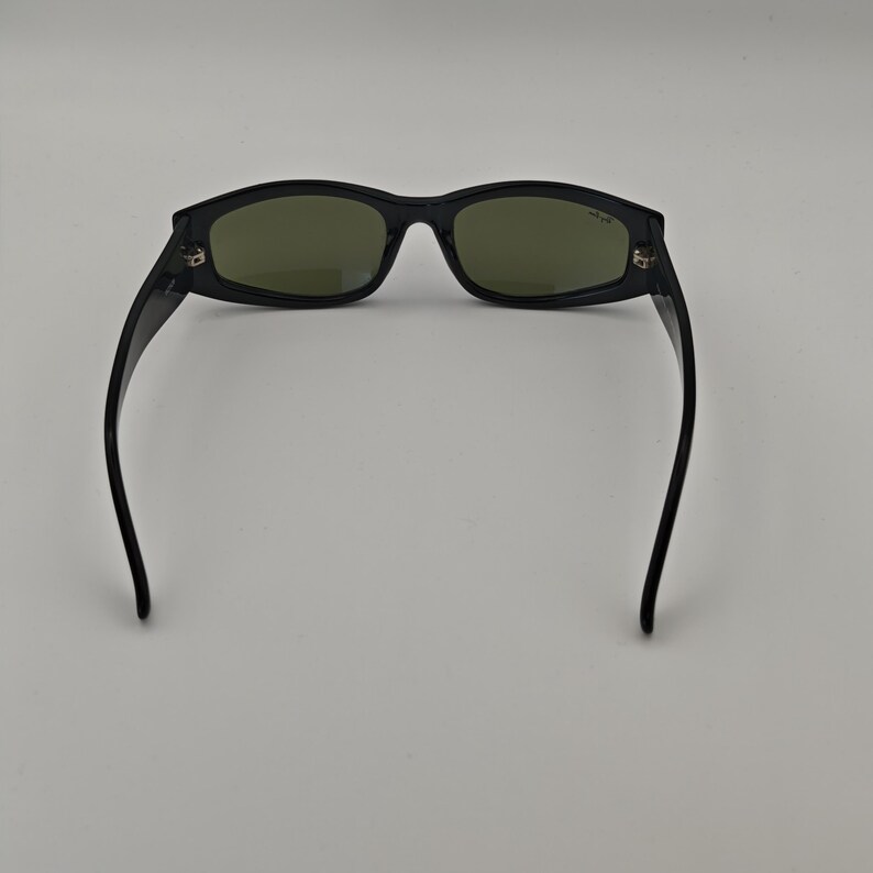 Może przedstawiać: Czarne okulary przeciwsłoneczne z prostokątnymi oprawkami i zielonymi szkłami. Okulary mają elegancki, nowoczesny design z lekko zakrzywioną oprawką i zausznikami. Szkła mają stonowany zielony kolor.