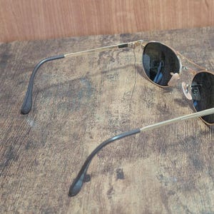May include: Gold-tone aviator sunglasses with dark lenses. The sunglasses have a double bridge and black temple tips. The lenses are round and reflect the surrounding environment. The frame is a shiny gold color.