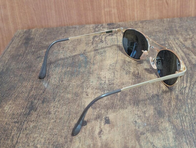 May include: Gold-tone aviator sunglasses with dark lenses. The sunglasses have a double bridge and black temple tips. The lenses are round and reflect the surrounding environment. The frame is a shiny gold color.