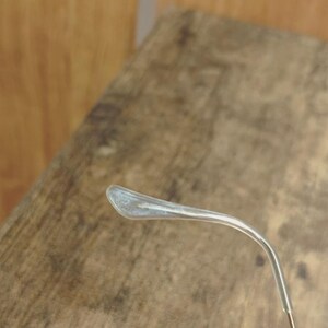 May include: Close-up of a clear eyeglass temple arm with a silver-colored tip. The arm curves gently, and the tip has a slightly frosted appearance. The background is a blurred wooden surface.