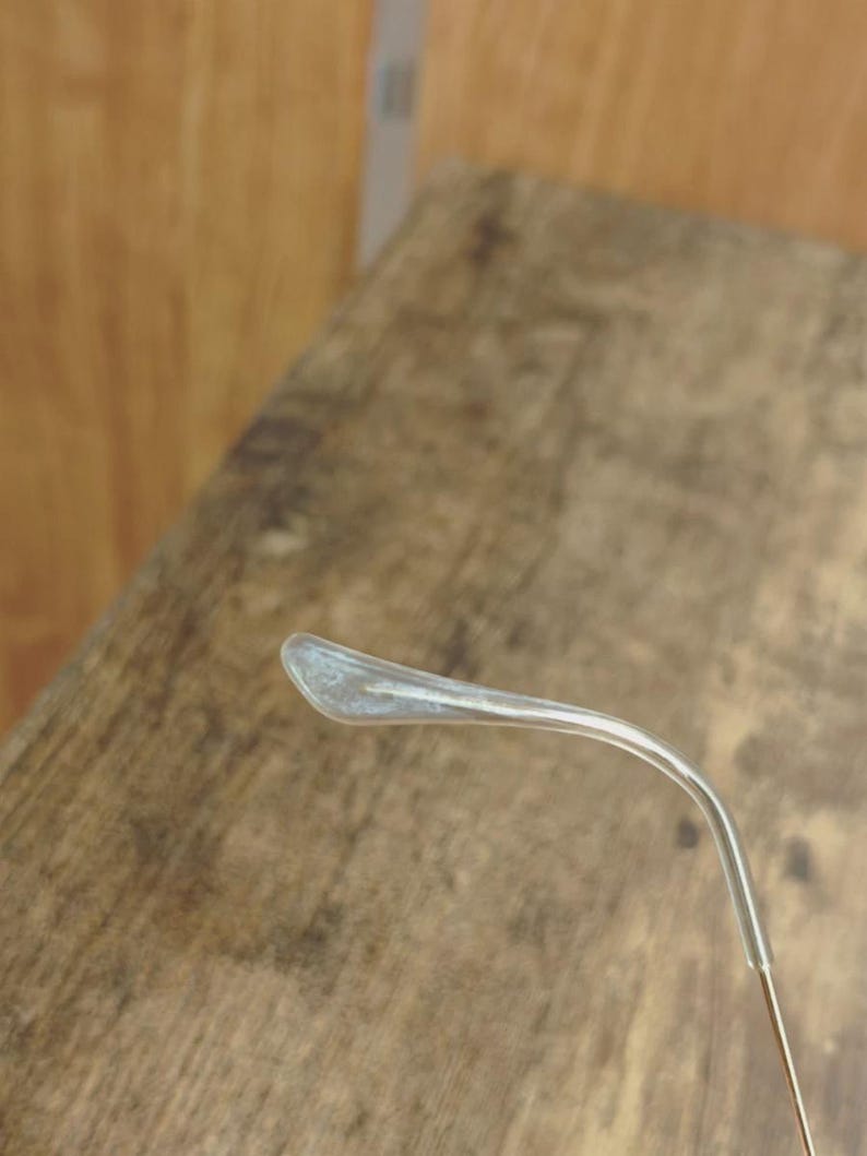 May include: Close-up of a clear eyeglass temple arm with a silver-colored tip. The arm curves gently, and the tip has a slightly frosted appearance. The background is a blurred wooden surface.